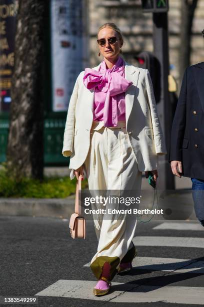 Guest wears black sunglasses, gold pendant earrings, a pink oversized knot neck shirt, a white blazer jacket, matching white large pants, a beige...