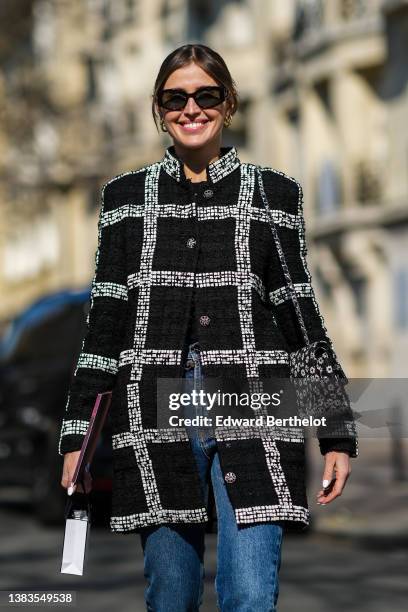 Darja Barannik wears black sunglasses, a black tweed with large white checkered print pattern buttoned coat from Chanel, a black with white flower...