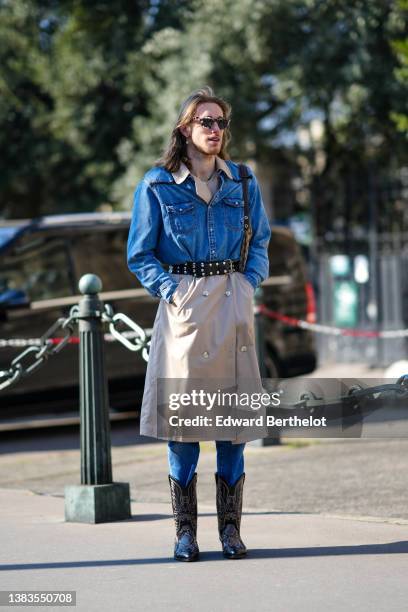 Guest wears sunglasses, gold pendant earrings, a gold necklace, a beige long buttoned trench coat, a blue denim jacket, a black shiny leather nailed...