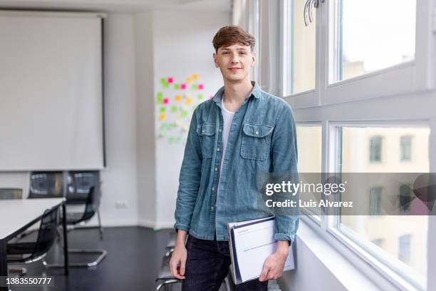 portrait of young man holding laptop standing by window at office - casual chic foto e immagini stock