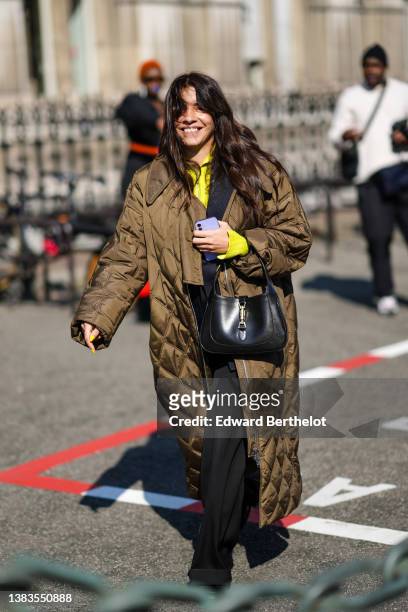 Guest wears a yellow zipper high neck pullover, a brown shiny quilted long oversized coat, a black shiny leather handbag, black suit pants, a black...