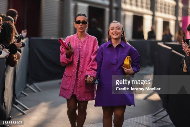 Fashion Week Guest is wearing a pink skirt, a matchy pink jacket and a small pink clutch, heels and sunglasses and an other woman is wearing a purple...