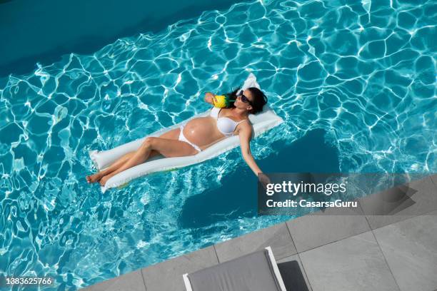 pregnant woman sunbathing on inflatable mat in a swimming pool - women in bathing suits stock pictures, royalty-free photos & images