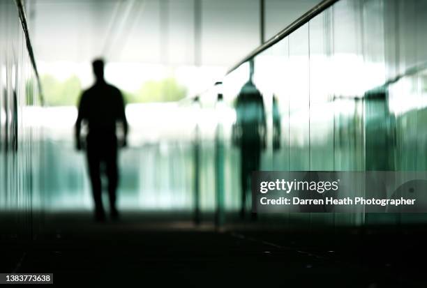 McLaren member of staff walking along an elevated walkway reflected in a glass balustrade inside the McLaren Technical Centre, Woking, Surrey, United...