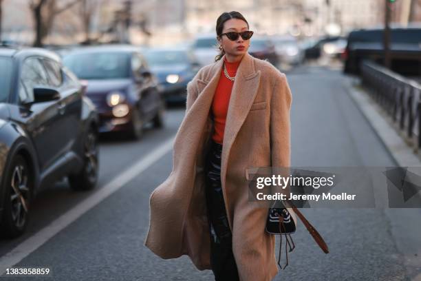 Fashion Week Gueswearing brown shades, a beige long coat, a red pullover, a gold chain, a black and white bag, black leather pants outside Sacai...