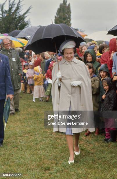 Queen Elizabeth ll, wearing a beige raincoat and a blue-and-white hat, holding a black umbrella, during a walkabout, New Zealand, 1st March 1977. The...