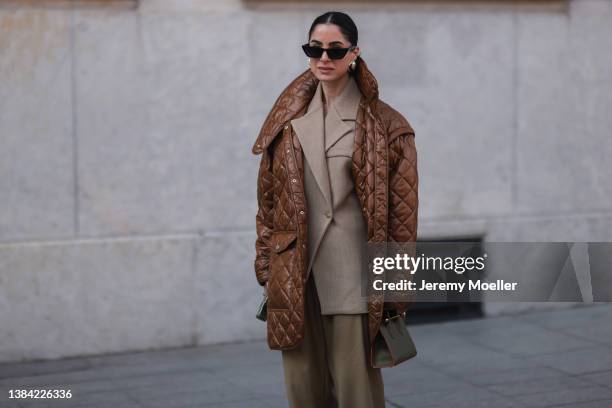 Guest wears black sunglasses, a beige blazer jacket, brown oversized puffy pants, a brown shiny quilted oversized puffer jacket, gold earrings, a...