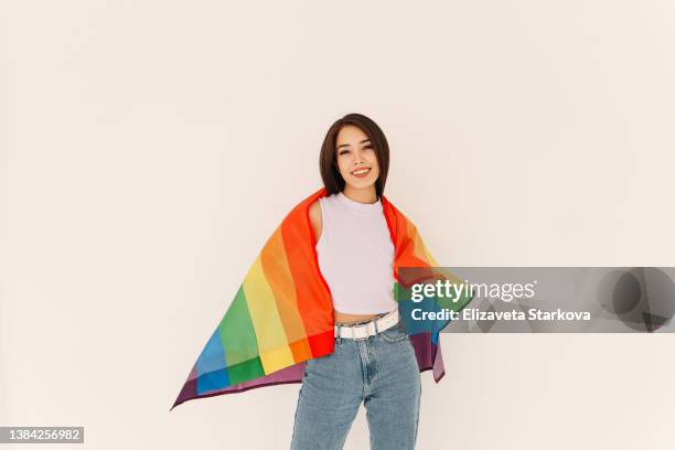 a happy smiling young woman a multiracial asian lesbian gay person stands rejoicing laughs and looks at the camera against the background of a multicolored rainbow flag an lgbt symbol - asian flags ストックフォトと画像