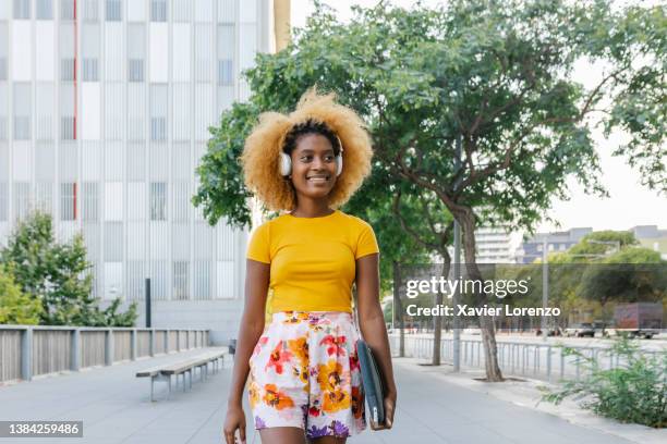 smiling afro woman listening to music with wireless headphones while walking down the street. - emerald city stock pictures, royalty-free photos & images