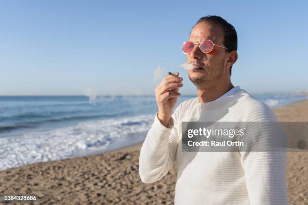 young man smoking marijuana joint cigarette, enjoying beautiful sunset at the seaside - cigarette bud stock pictures, royalty-free photos & images