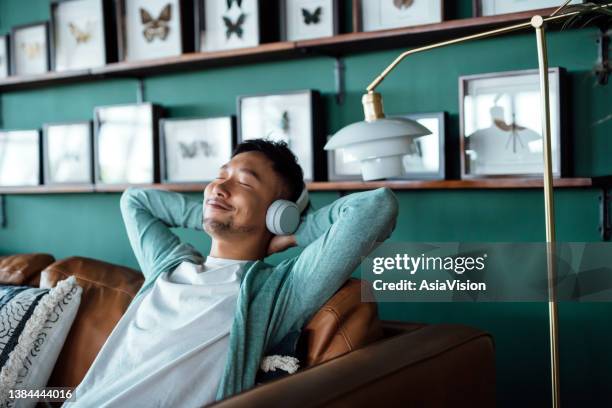 jeune homme asiatique avec les mains derrière la tête, se relaxant sur le canapé et écoutant de la musique avec des écouteurs à la maison. jeune homme détendu allongé sur un canapé avec de la musique. style de vie relaxant, personnes et concept te - être étendu photos et images de collection
