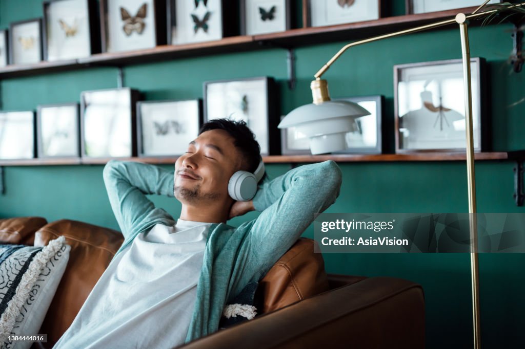 Young Asian man with hands behind head, relaxing on sofa and listening to music with headphones at home. Relaxed young man lying on sofa with music. Relaxing lifestyle, people and technology concept