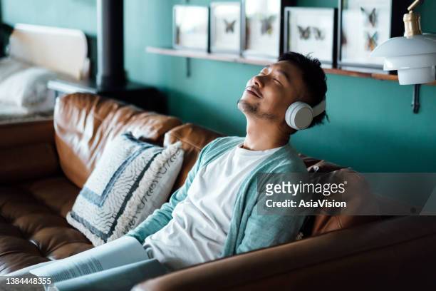 Young Asian man listening to music with headphones and reading book in living room at home. Relaxed young man with eyes closed lying on sofa with music while reading book. Relaxing lifestyle, people and technology concept