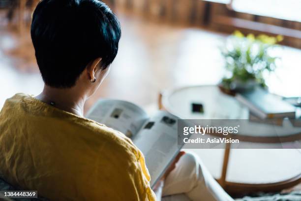 over the shoulder, rear view of senior asian woman sitting on sofa in the living room, relaxing and reading book at home. relaxing lifestyle, people and retirement concept - escapism reading stock pictures, royalty-free photos & images
