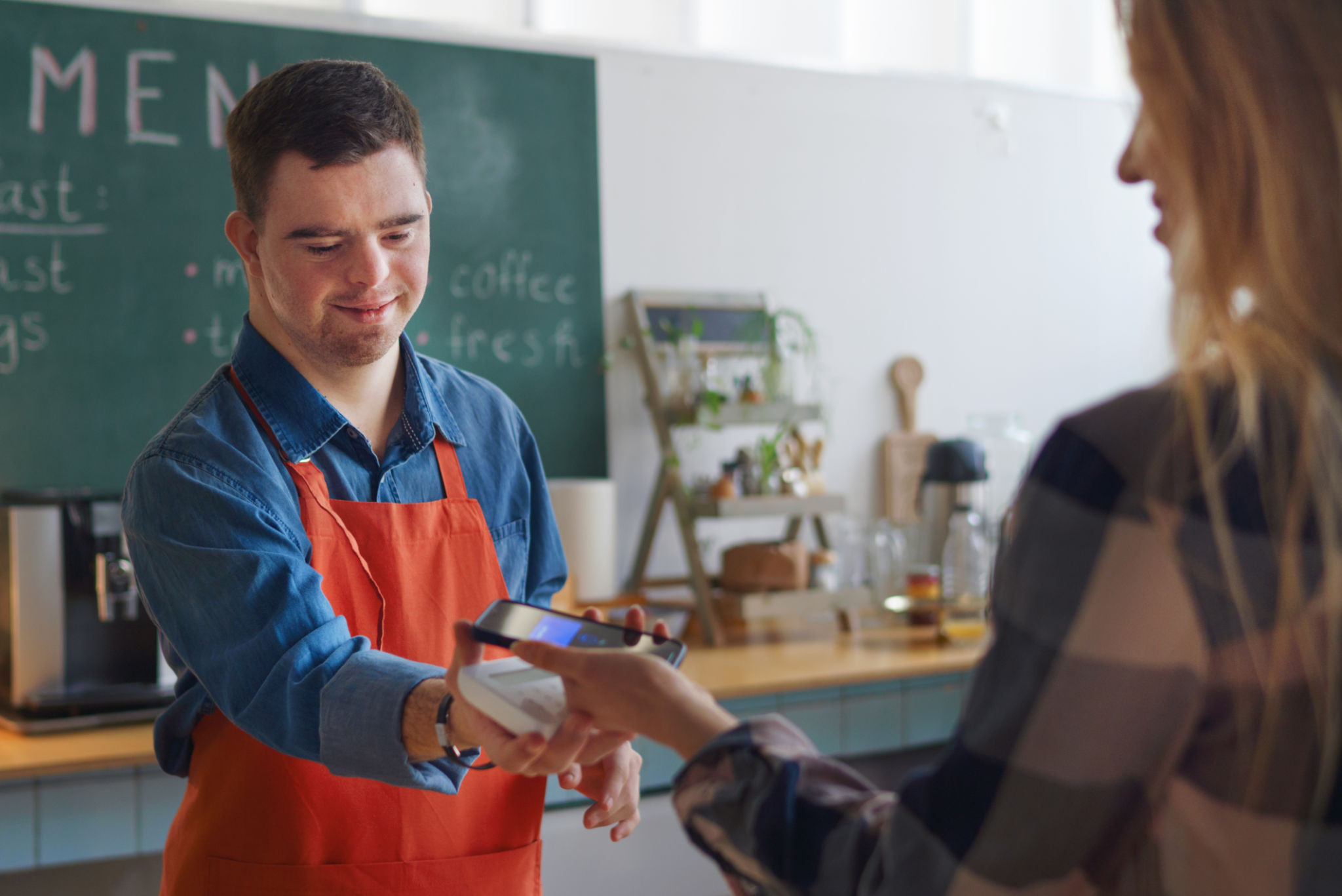Cheerful young Down Syndrome waiter taking contactless smartphone payment from costumer in take away restaurant. Cheerful young Down Syndrome waiter taking contactless smartphone payment from costumer in take away restaurant.