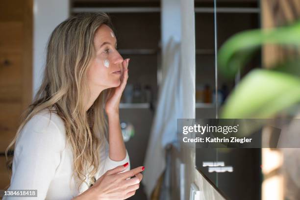 woman applying face cream in front of bathroom mirror - cream coloured stock pictures, royalty-free photos & images