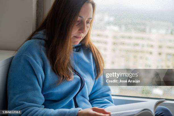 mujer leyendo un libro junto a la ventana - descontrair fotografías e imágenes de stock