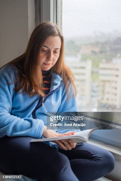 mujer leyendo un libro junto a la ventana - descontrair fotografías e imágenes de stock