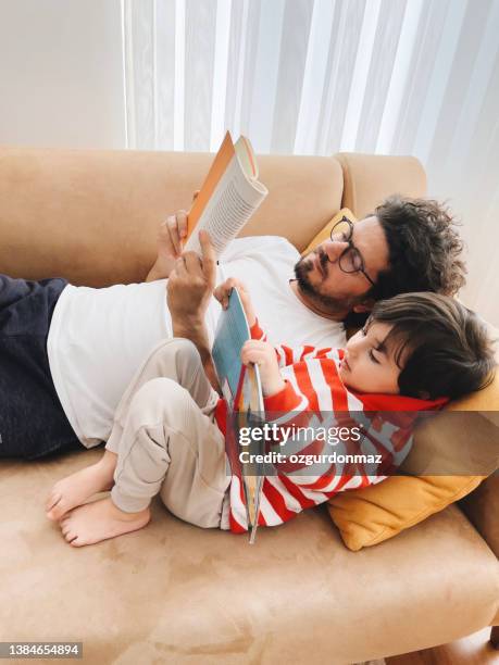 father and son lying on sofa at home and reading book together - kids read stockfoto's en -beelden