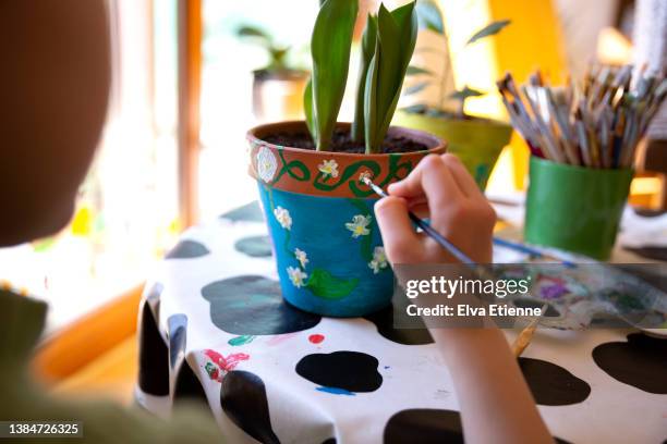 over the shoulder view of a child painting a terracotta plant pot with bright coloured paints on a dining table in springtime. - pintar maceta fotografías e imágenes de stock