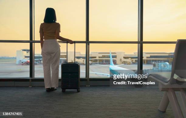 travel tourist standing with luggage watching sunset at airport window. woman looking at lounge looking at airplanes while waiting at boarding gate before departure. travel lifestyle. transport and travel concept - luggage stockfoto's en -beelden