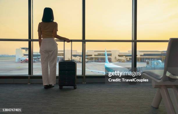 travel tourist standing with luggage watching sunset at airport window. woman looking at lounge looking at airplanes while waiting at boarding gate before departure. travel lifestyle. transport and travel concept - carry on bag stock pictures, royalty-free photos & images