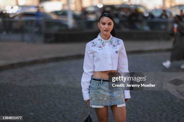 Camila Mendes seen wearing gold earrings, a white flower embroidered cropped shirt from Miu Miu, a blue denim short skirt with flower details, a...