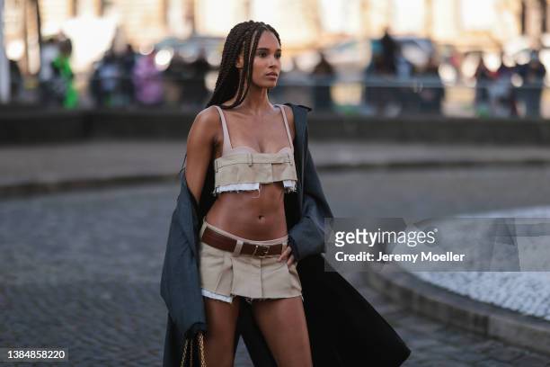 Model Cindy Bruna seen wearing a Miu Miu beige bra crop top with a matching beige pleated mini skirt, a brown leather belt, a grey coat, knee-high...