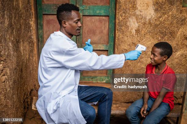 männlicher afrikanischer arzt, der die temperatur eines jungen misst, abgelegenes dorf in ostafrika - doctors without borders stock-fotos und bilder