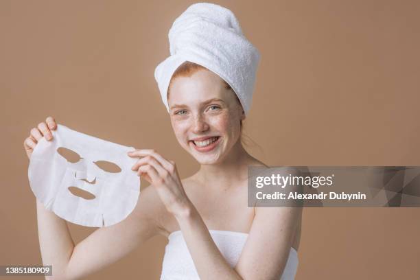 happy young caucasian woman taking care of facial skin with lotion and sheet mask holding in hands with copy space. smiling emotional girl concept of beauty and healthy hygiene - cloth mask stock-fotos und bilder