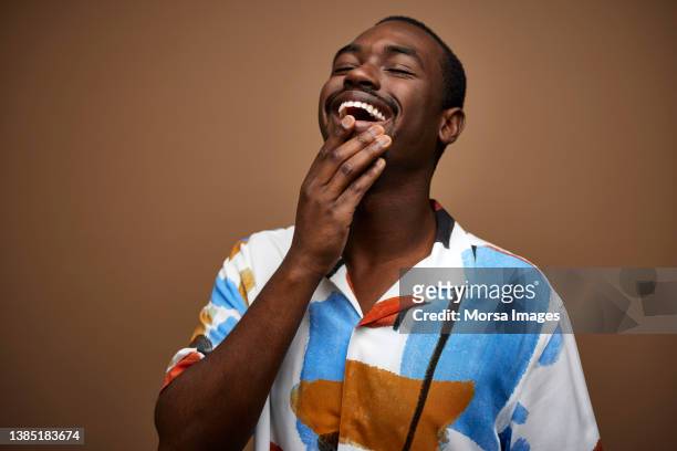 cheerful young african man with hand on chin against brown background - schwarzes hemd stock-fotos und bilder