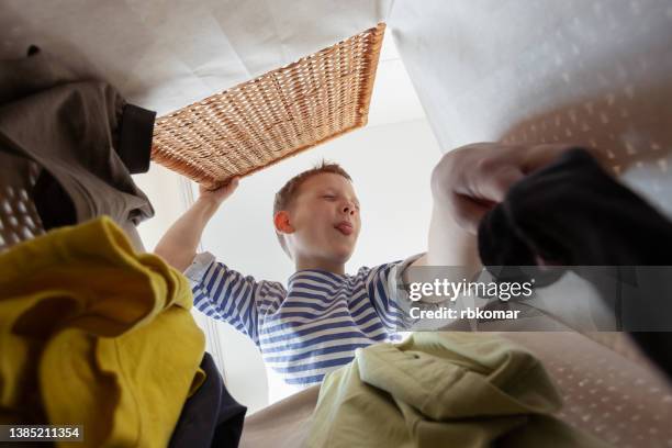 little boy squinting from an unpleasant smell throws dirty clothes into a basket, bottom view from the inside. disgust of children in housework - boy in a box stockfoto's en -beelden