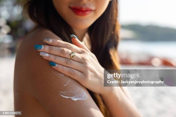 beautiful happy cute woman applying suntan cream to her shoulder with beach background - gebruind stockfoto's en -beelden