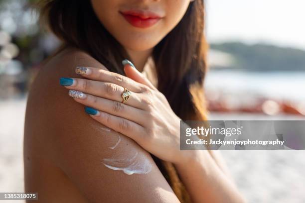 beautiful happy cute woman applying suntan cream to her shoulder with beach background - proteção uv - fotografias e filmes do acervo