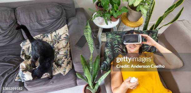 latino-frau in ihren dreißigern mit lockigem haar und gelber bluse mit virtual-reality-brille, die auf einem sofa in einem wohnzimmer mit pflanzen und einer schwarzen katze liegt - cat vr stock-fotos und bilder