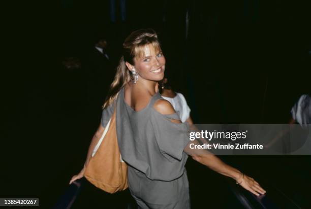 America fashion model Christie Brinkley, wearing a grey outfit, her shoulder exposed through slits in the sleeve, a tan bag over her shoulder, circa...