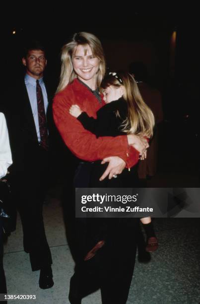 America fashion model Christie Brinkley, wearing black trousers and a red jacket, carrying her daughter Alexa Ray Joel at an event, circa 1990.