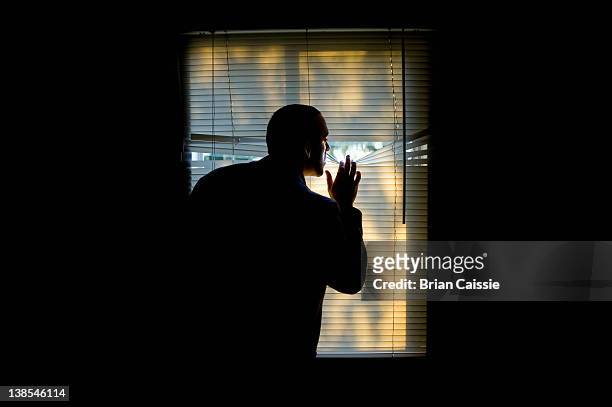 a man peeking outdoors through the blinds of a darkened room - peep window stock-fotos und bilder