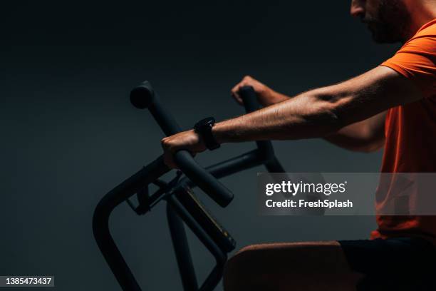 a side view of an unrecognizable male athlete in orange t-shirt riding an exercise bike at the gym - peloton app stock pictures, royalty-free photos & images