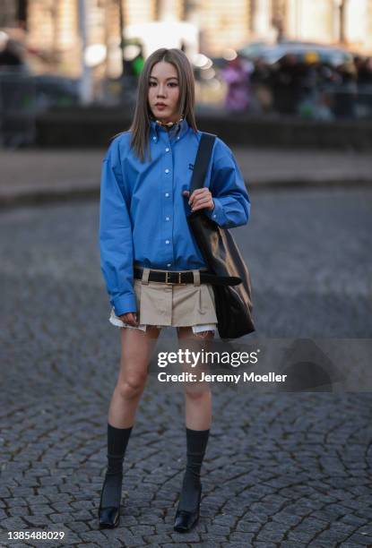 Jenny Suet In Tsang seen wearing a blue oversized shirt from Miu Miu, a gold and pearls necklace, a black shiny leather large shoulder bag from Miu...