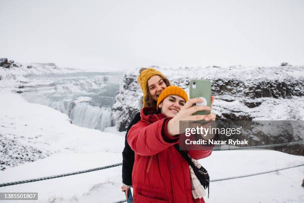 two cheerful women doing selfies at the gullfoss waterfall background - frozen waterfall stockfoto's en -beelden