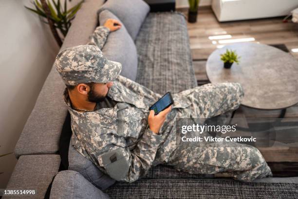 soldier sitting on sofa in living room and using a smartphone - rang stockfoto's en -beelden