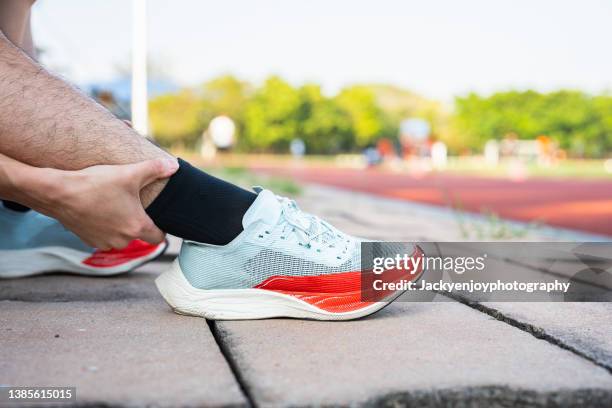 low section of man holding aching ankle while kneeling outdoors - sehnenscheidenentzündung stock-fotos und bilder