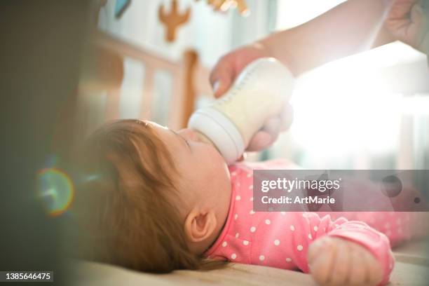 cute happy baby girl in pink bodysuit drinking milk from baby bottle indoors in a sunny morning - happy mobile stockfoto's en -beelden