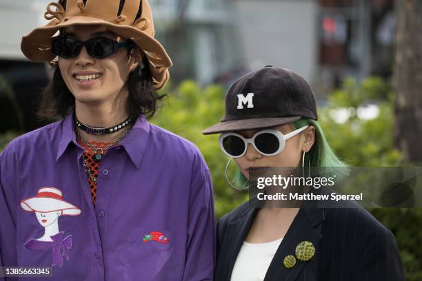 Guests are seen wearing purple collared shirt, orange mesh shirt, brown bag, navy pants with scarf knee accessory, black shoes and brown felt hat;...