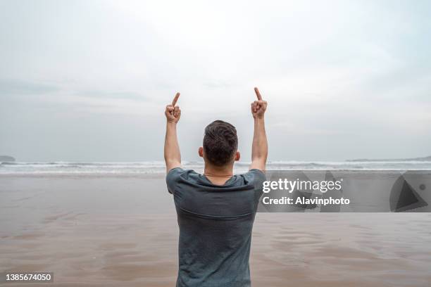 young man making offensive symbol with his hands on the sea shore at the beach having fun - global mindset stock pictures, royalty-free photos & images