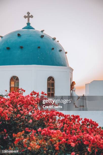 young woman enjoys travelling and looking at the sunset view in the front of a famous traditional blue dome church and red flowers in santorini island, greece thire village, greece - mediterranean climate stock pictures, royalty-free photos & images