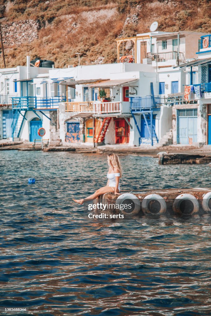Young woman slapping feet in water and looking at view of traditional fisherman village Klima on Milos island, Greece