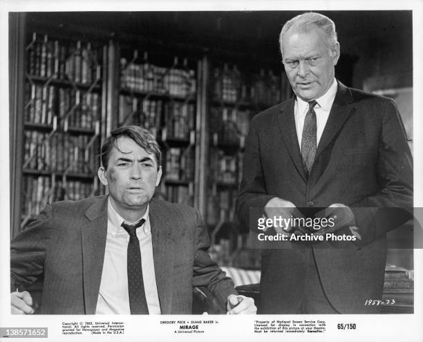 Gregory Peck sitting looking disheveled in a suit an unknown actor is standing behind him in a scene from the film 'Mirage', 1965.