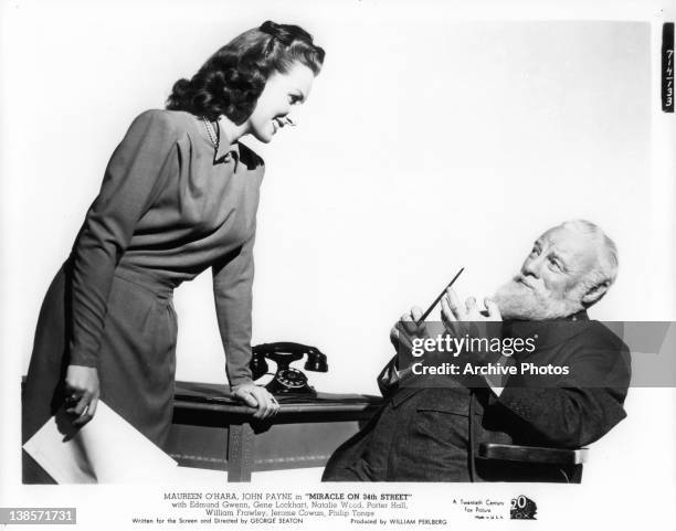 Maureen O'Hara bending down talking with Edmund Gwenn sitting in a chair who has a full beard in a scene from the film 'Miracle On 34th Street', 1947.